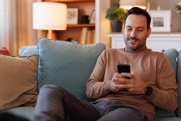 Portrait of confident handsome man text messaging over smart phone while relaxing on sofa in home office