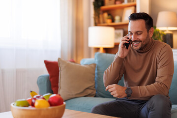 Fototapeta premium Portrait of handsome man smiling and talking over smart phone while sitting on sofa in living room