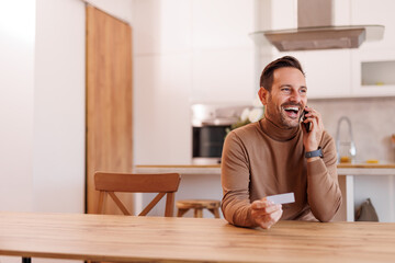 Mid adult man with credit card talking over smart phone while sitting at wooden dining table at home