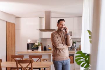 Smiling mid adult man looking through window and talking over smartphone while standing by dining...