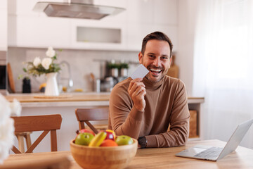 Portrait of confident mid adult man showing credit card while sitting with laptop at dining table at home
