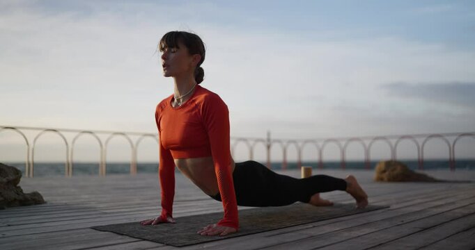 A confident brunette yoga master girl in an orange sweater performs a cobra pose during her yoga class on a special carpet on a beach covered with boards at Sunrise in summer