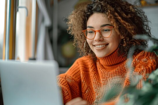 Happy woman wearing glasses and orange sweater using laptop sitting at desk taking notes watching online webinar in daylight from window remote working studying learning digital course