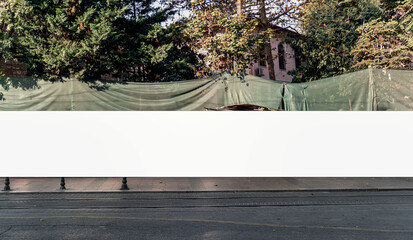 White banner stretched across construction site with green mesh fencing, trees and sidewalk in urban setting, ideal for signage or advertising mockups © Bonsales