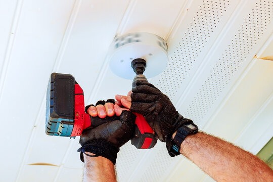 An individual wearing gloves holds cordless drill that has hole saw attachment attached, cuts circular hole in white ceiling to accommodate new light fixture