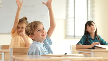 Caucasian boy raising hand for asking and answering teacher surrounded by diverse children studying at classroom. Happy student working together, voting, volunteering, calling instructor. Pedagogy.