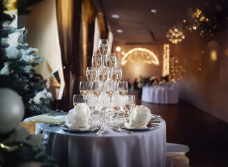 A champagne pyramid sits on a table at a wedding reception.  The pyramid is in the foreground with white table cloths, plates, and silverware.  The background is blurred and shows a wedding ceremony t