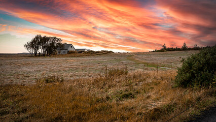 Nordic farm house with outbuildings is nestled into a small grove of trees at the far side of a grassy field showing Fall colors. It's twilight, and the clouds reflect shades of orange and yello.