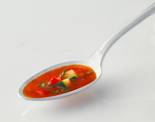 A single spoon loaded with vibrant Tomato soup on a white surface. showcasing diced vegetables like peppers and zucchini in a rich broth. Isolated on white