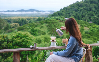 Rear view of a woman making drip coffee while sitting on wooden balcony with a cat and a beautiful...