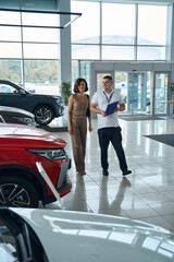 Salesman manager with file folder and female customer standing in car showroom