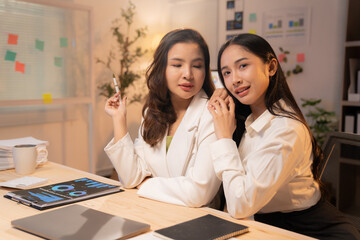 Two asian businesswomen are working together on a project in a modern office at night, discussing strategy and analyzing financial data with charts and graphs, demonstrating teamwork and dedication