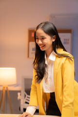 Young asian businesswoman wearing yellow jacket and white shirt is smiling and leaning on desk in...