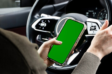 Close-up of woman's hands holding smartphone with green empty copy space on screen mockup behind the wheel of sport's car rear view, connecting phone to a electric car.