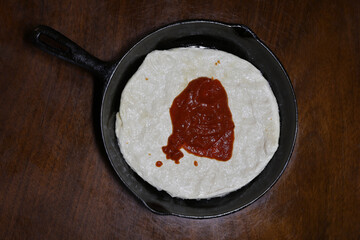small pizza dough with a spoonful of sauce, pressed into a cast iron pan placed on a dark wood...