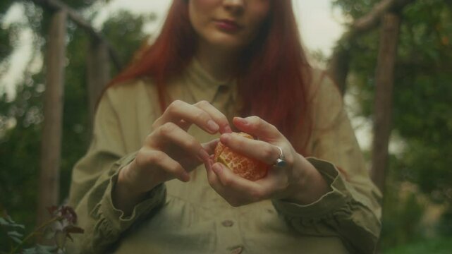 A ginger young woman sits on a wooden bridge in an orange orchard peeling an orange