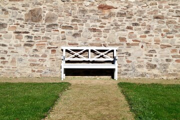 White wooden bench standing in front of an old, solid stone wall. The bench has a simple design and...