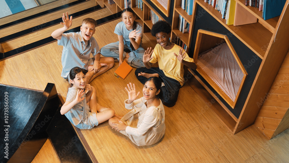 Poster top view of diverse children sitting in circle while looking at camera and waving hands at library s