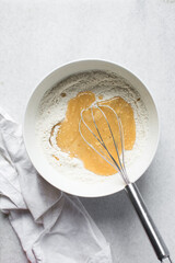 Overhead view of vanilla cake batter being whisked in a white ceramic bowl, flatlay of cake batter being mixed, process of making cake
