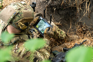 A soldier in tactical gear operates a control tablet in the field, using technology for reconnaissance with an aerial view display on the screen.