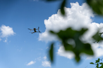 A drone flies against a bright blue sky with scattered clouds, surrounded by blurred green leaves...
