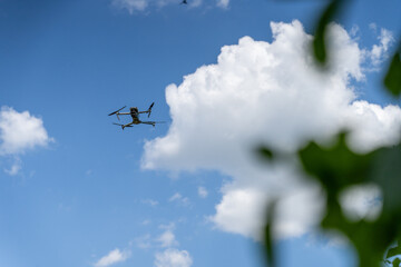 A drone flies against a bright blue sky with scattered clouds, surrounded by blurred green leaves in the foreground. aerial reconnaissance work