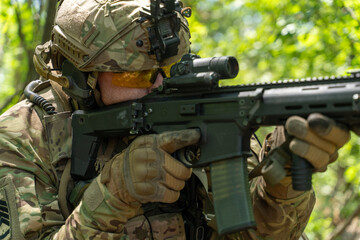 A soldier in full camouflage gear takes aim with a rifle in a wooded area, showcasing military equipment and focus in a natural setting. military operations in the forest