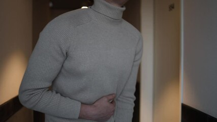A person in a beige turtleneck leans forward, clutching their abdomen, showing signs of bloating and intestinal discomfort. The subdued hallway lighting highlights a sense of vulnerability and unease.