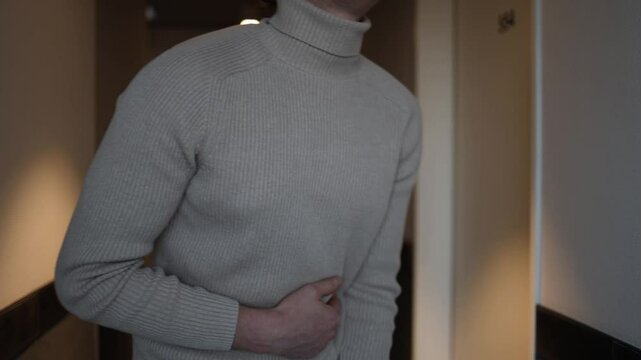 A person in a beige turtleneck leans forward, clutching their abdomen, showing signs of bloating and intestinal discomfort. The subdued hallway lighting highlights a sense of vulnerability and unease.