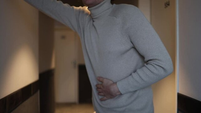 A person in a beige turtleneck clutches their stomach, appearing nauseous and distressed, in a dimly lit hallway. The subdued lighting enhances the sense of discomfort and vulnerability in the scene.