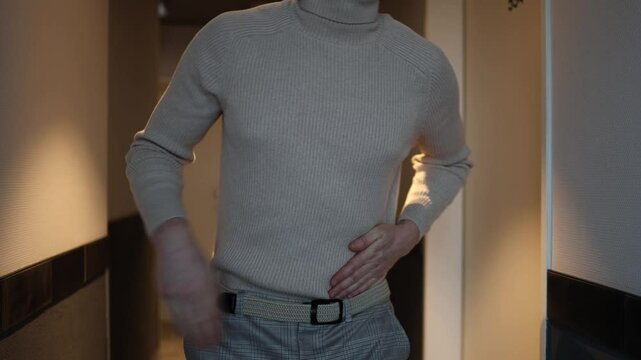 A person in a beige turtleneck and plaid trousers stands in a hallway, clutching their upper abdomen with a pained expression, suggesting discomfort in the pancreas area. The dim lighting adds a sombe