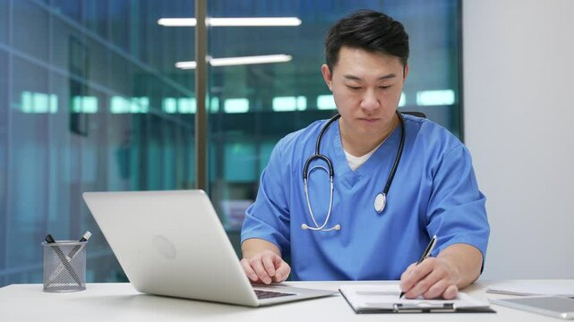 Asian male doctor in office, focused on writing and using laptop computer. Medical worker physician takes notes or is busy with paper work sitting at desk at the workplace in a modern hospital clinic