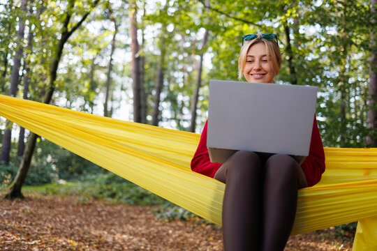 Cheerful young woman with laptop sitting on hammock in forest - Powered by Adobe