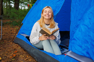 Smiling happy woman with a book in her hands sits in a tent surrounded by forest