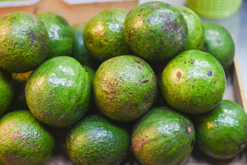 The avocados are green with some brown spots and rough patches on their skin. They are stacked closely together, likely at a market or store display.