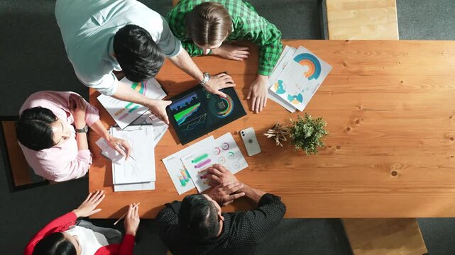 Top view of business man using tablet display increasing graph while diverse marketing people sharing idea or planing strategy at meeting table with stock market statistic scatter around. Convocation.