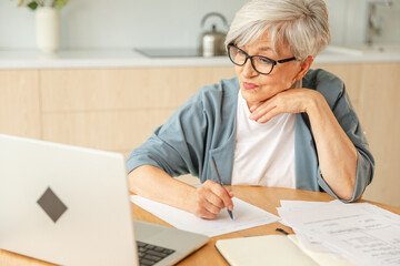 Middle aged senior woman using laptop computer writing notes at home. Focused mature old businesswoman work in home office making written records doing online work. Mature boss professional worker
