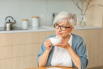 Good morning. Comfort relaxed senior old elderly woman grandmother drinking hot beverage tea coffee in kitchen at home. Mature woman resting enjoy calm morning at modern home alone. Pension concept