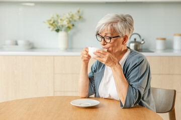 Good morning. Comfort relaxed senior old elderly woman grandmother drinking hot beverage tea coffee in kitchen at home. Mature woman resting enjoy calm morning at modern home alone. Pension concept