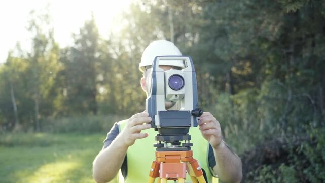 A man is holding a surveying instrument in a field. The instrument is a total station, which is used to measure distances and angles. The man is wearing a safety helmet and a green vest