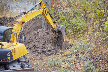 An excavator pouring out excavated soil from its bucket, close-up of excavator work at a construction site in a wooded area.