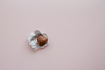 A heart-shaped chocolate candy in a red wrapper unwrapped on a pink background, sweets for...