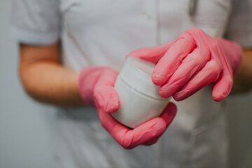 A nurse in a white coat and pink gloves holds a white jar of cream for wounds and scratches on skin