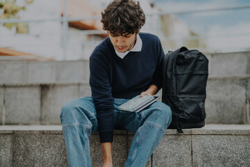 A young male student sits on outdoor steps in the city, focused on his studies with a notebook and backpack nearby. The casual setting reflects a relaxed yet productive atmosphere.