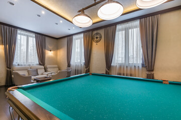 A luxurious, well-lit billiard room, with windows. Cozy beige sofas at a small table.