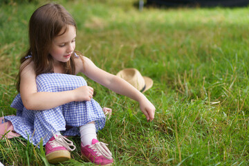 Fototapeta premium Little cute girl in a sundress and a straw hat sits on the lawn. The concept of children's health, education, fun