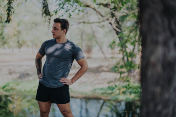 Fototapeta premium A young man stands casually in a park, enjoying the peaceful outdoor setting. He appears relaxed and contemplative, surrounded by greenery and natural beauty on a sunny day.