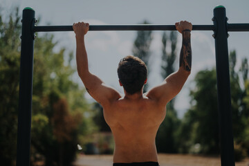 Naklejka premium Rear view of a shirtless young man performing pull-ups on a bar in a park, showcasing fitness, strength, and a healthy outdoor lifestyle.