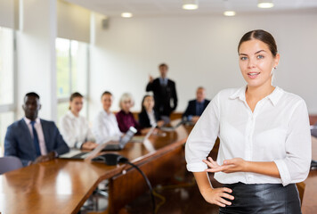 Smiling millennial white dark-haired businesswoman in white shirt standing in conference room during group meeting posing with hand palm up thus showing welcoming or presenting hand gesture