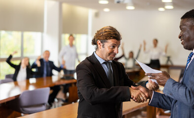 Imposing middle aged white boss welcoming successful african male candidate congratulating him on getting hired at new job in international company in front of other company employees watching them